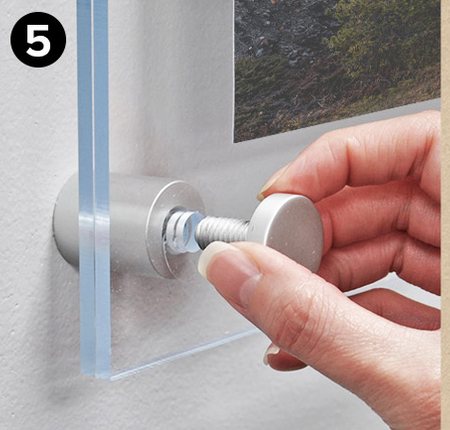 A hand tightening a silver standoff cap on a mounted acrylic frame